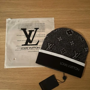 Svart mössa - Svart och grå mössa med ikoniskt monogrammönster och tydlig logga framtill. Mössan har en vit rand och är tillverkad i mjukt material som känns lyxigt. Perfekt accessoar för att lyfta din stil.