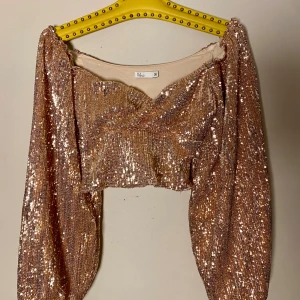 Glittrig croppad topp från NLY Trend - Säljer en glittrig croppad topp från NLY Trend i guld med paljetter över hela ytan. Toppen har långärmade puffärmar, omlott framtill och är tillverkad i 100% polyester. Perfekt för att sticka ut med extra shimmer.