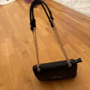 Svart quiltad axelväska från Valentino - Stilren svart axelväska från Valentino med quiltat mönster och guldfärgade kedjor. Väskan är tillverkad i skinnimitation och har ett lock med magnetlås. Perfekt storlek för det viktigaste och snygg detalj till din outfit. Öppen för pris förslag men denna är använd endast fåtal gånger så säljer ej för ”billigt” 