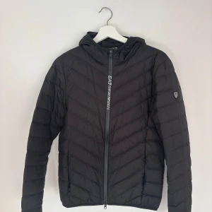 Svart pufferjacka från EA7 Emporio Armani - Svart pufferjacka från EA7 Emporio Armani med huva och snyggt mönstrad dragkedja framtill. Jackan har quiltad design, lång ärm och logotyp på vänster ärm. 