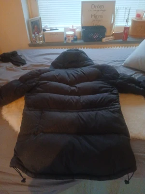 Svart dunjacka med huva från boomarang  - Säljer en svart, fluffig dunjacka från Downemong med huva och snörning nertill. Jackan har både dragkedja och knappar framtill, samt breda quiltade sektioner som ger en cool och chunky look. Perfekt för kalla dagar när du vill hålla värmen och se chill ut.