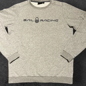 Grå Sail Racing sweatshirt - Snygg grå sweatshirt från Sail Racing med svart logotyp tryckt på bröstet. Tröjan har rund hals, ribbade muddar och är långärmad. Perfekt för dig som gillar stilren och sportig design. Mjuk insida och klassisk passform.