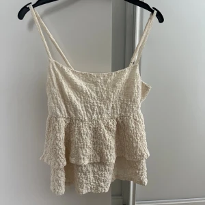 Beige volang linnetopp med smock - Säljer en ljusbeige linnetopp med smala axelband och dubbla volanger. Toppen har smockad struktur som ger en cool och avslappnad vibe. Perfekt för varma dagar och enkel att matcha med jeans eller kjol.