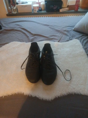 Svarta fotbollsskor med snörning - Säljer ett par svarta fotbollsskor med klassisk rund tå och snörning. Skorna har en stilren look med diskret mönster på ovandelen och platt sula. Perfekta för dig som vill ha en clean och sportig stil på planen.