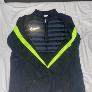 Nike Therma-Fit svart tröja XL - Nike Therma-Fit tröja i svart med quiltad front och halv dragkedja. Snygga neongula detaljer längs ärmarna och bröstet, samt Nike-logga på vänster sida. Materialet är polyester och syntet, perfekt för träning och kyligare dagar.
