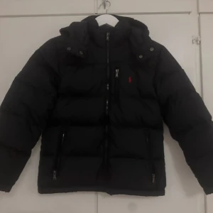 Svart pufferjacka från Polo Ralph Lauren - Svart pufferjacka från Polo Ralph Lauren med huva, dragkedja och två sidofickor med zip. Jackan har det klassiska röda Polo-logot på bröstet och quiltad design. Perfekt för kalla dagar och har en clean, sportig vibe. om man vill hänga det går inte strött är sönder! 