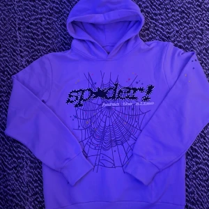 Lila hoodie från Spider med tryck - Lila hoodie från Spider med coolt spindelnäts- och stjärntryck på bröstet. Framsidan har texten 'Spider! heres the slime' i svart och vitt med prickar. Tröjan har huva och långa ärmar, tillverkad i 100% bomull. Perfekt för dig som gillar unika prints och färg.
