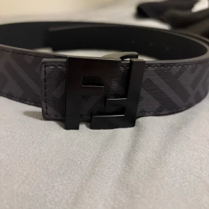 Svart Fendi bälte med FF-spänne - Säljer ett stilrent svart Fendi bälte med det ikoniska FF-spännet i matt svart metall. Bältet har ett diskret mönster med Fendi-loggor längs hela bandet och är tillverkat i skinn. Perfekt accessoar för att lyfta din outfit med en lyxig touch.