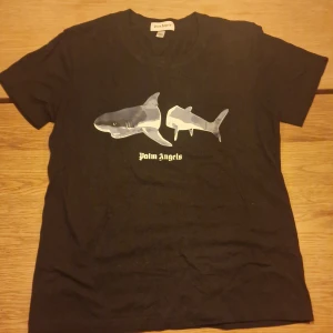 Svart Palm Angels t-shirt med hajtryck - Svart t-shirt från Palm Angels med ett unikt hajmotiv i grå och vita toner på bröstet. Klassisk rund hals och korta ärmar. Materialet är mjuk bomull som känns skönt mot huden. Palm Angels-loggan är tryckt under motivet för extra streetkänsla.