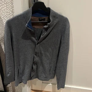 Grå cardigan från Massimo Dutti - Snygg grå kofta från Massimo Dutti i mjuk bomull och kashmirmix. Koftan har ribbad krage, knappar framtill och två fickor. Perfekt för lager-på-lager och chill stil. Passar dig som gillar klassiska plagg med modern touch.