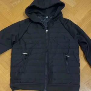 Svart hybridjacka  - Svart hybridjacka med huva och quiltad framsida. Jackan har två sidofickor med dragkedja och hel dragkedja framtill. Den lätt och skön. Perfekt för kyliga dagar när du vill ha en stilren look. Jackan är i storlek S men kan även passa M. Priset kan diskuteras!