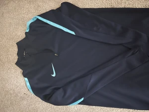 Nike Dri-Fit mörkblå half zip tröja - Nike Dri-Fit tröja i mörkblått med ljusblå detaljer längs axlar och ärmar. Tröjan har lång ärm, half zip framtill och ett stilrent Nike-logotryck på bröstet. Perfekt för träning eller sportiga dagar, tillverkad i lätt polyester som andas.