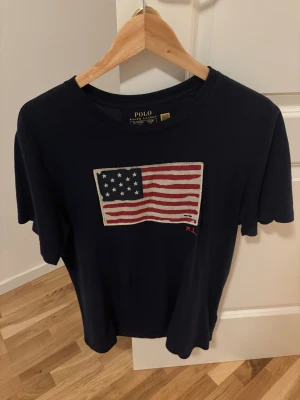 Mörkblå t-shirt Polo Ralph Lauren XL - Snygg mörkblå t-shirt från Polo Ralph Lauren i storlek XL med broderad amerikansk flagga på bröstet och RL-detalj i rött. Klassisk rund hals och korta ärmar. Tillverkad i mjuk bomull som känns skön mot huden.