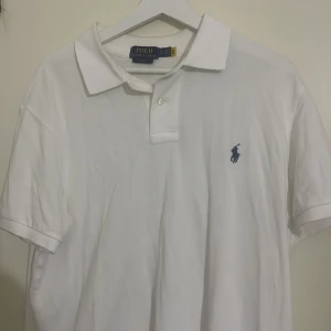Vit Polo Ralph Lauren piké XL - Vit pikétröja från Polo Ralph Lauren i slim fit med klassisk krage och två knappar. Ikonisk blå broderad logga på bröstet. Tillverkad i bomull som känns skön mot huden. Perfekt för en clean och stilren look. Tröjan passar L och har använts fåltal gånger, nypris 1595 kr