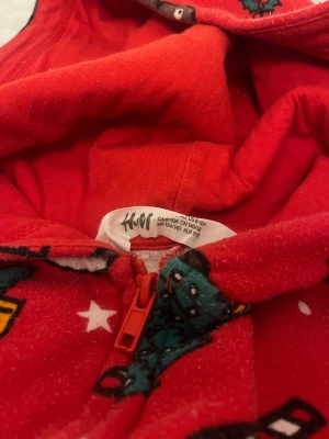 Röd julmönstrad jumpsuit från H&M - Supermysig röd jumpsuit med huva från H&M, täckt av juliga motiv som pepparkakor, snögubbar, julgranar och polkagrisar. Mjuk fleece-känsla och dragkedja framtill gör den enkel att ta på. Perfekt för chilliga vinterkvällar!