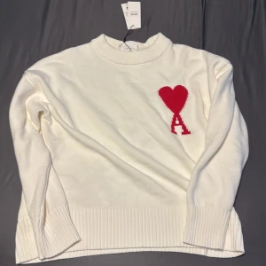 Vit stickad tröja från Ami Paris - Vit stickad tröja från Ami Paris med röd hjärtlogga på bröstet. Tröjan har ribbade muddar vid ärmslut och nederkant samt rund halsringning. Mjuk och skön bomullskvalitet, perfekt för dig som gillar stilrena detaljer med en twist.