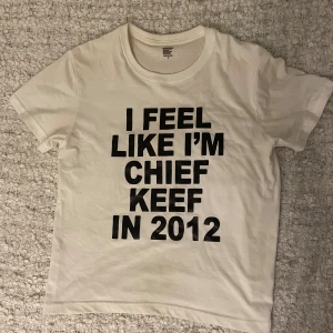 Vit t-shirt med Chief Keef-tryck - Vit t-shirt i bomull med stort svart texttryck på framsidan: 'I FEEL LIKE I'M CHIEF KEEF IN 2012'. Klassisk passform med rund hals och korta ärmar. Perfekt statement-plagg för dig som gillar hiphop och vill sticka ut.