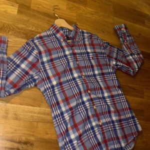 Rutig skjorta från Ralph Lauren - Riktigt snygg rutig skjorta från Ralph Lauren i blått, rött och vitt. Skjortan har klassisk button-down krage, broderad logga på bröstet och är tillverkad i stretchig bomull. Perfekt passform med slim fit och långa ärmar. Stängs med knappar hela vägen. Pris är ej hugget i sten!🪨 