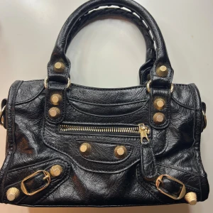 Svart handväska med gulddetaljer - Cool svart handväska i skinn med snygga guldfärgade nitar och spännen. Väskan har flätade handtag, avtagbar axelrem och flera dragkedjefack. Perfekt storlek för det viktigaste och ger en edgy vibe till din outfit.
