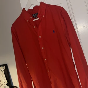 Röd skjorta från Polo Ralph Lauren - Riktigt snygg röd skjorta från Polo Ralph Lauren i slim fit. Klassisk button-down krage, vita knappar och blå broderad logga på bröstet. Skjortan är långärmad och gjord i mjuk bomull, perfekt för en snygg koll nu i juletiderna. Tveka inte med att skriva, priset är ej hugget i sten!🪨 