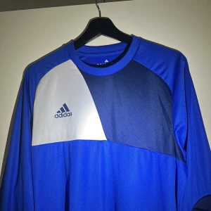 Blå långärmad Adidas målvaktströja - Säljer en blå långärmad Adidas målvaktströja med vita och mörkblå detaljer. Tröjan har vadderade armbågar och är gjord i lätt, ventilerande Climalite-material som håller dig torr på planen. Adidas-logga på bröstet och snygg sportig design.