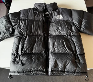 Svart North Face 1996 Retro  - Svart pufferjacka från The North Face i storlek M. Jackan har en hög krage, två sidofickor med dragkedja och en innerficka med zip. Materialet är syntet och dun, perfekt för kalla dagar. Klassisk logga på bröstet och insidan, även på baksidan! 