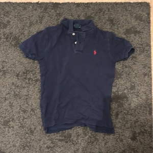 Marinblå pikétröja Polo Ralph Lauren - Snygg marinblå pikétröja från Polo Ralph Lauren i bomull. Klassisk krage, korta ärmar och två knappar framtill. Röd broderad logga på bröstet ger en stilren touch. Perfekt för dig som gillar tidlös och sportig stil.