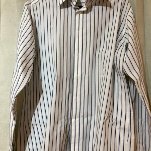 Randig skjorta från Selected, vit/blå- NY - Vit skjorta från Selected med smala blå ränder, klassisk krage och knäppning framtill. Långärmad modell i mjuk bomull, perfekt för en clean och stilren look. Passar dig som gillar enkel men snygg design.