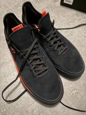 Svarta Adidas sneakers med orange detaljer - Snygga svarta Adidas sneakers med orange logga och detaljer på sidan samt plös. Aldrig använda. Ovandel i mocka med klassisk snörning och platt sula. Yttersulan har ett coolt hexagonmönster i orange och svart som ger extra grepp. Perfekt för dig som gillar stilrena och sportiga skor. Lådan finns kvar också. 