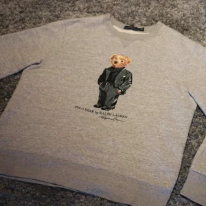 Grå Polo Bear - Grå sweatshirt från Polo Ralph Lauren med ikoniskt Polo Bear-tryck framtill. Tröjan har rund hals, långa ärmar och är tillverkad i mjuk bomullsmix. Perfekt för dig som gillar streetstyle och vill ha något klassiskt men ändå lekfullt.
