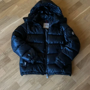 Svart pufferjacka från Moncler - Säljer en svart pufferjacka från Moncler, modell Maya Giubbotto. Jackan har huva, dragkedja framtill och Moncler-logga på ärmen. Den är tillverkad i blankt syntetmaterial och har en quiltad design som ger en schysst volym. Perfekt för kalla dagar och riktigt snygg streetstil.