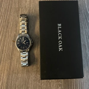 Silverfärgad klocka från Black Oak - Stilren klocka från Black Oak med silverfärgat metallarmband och mörkblå urtavla. Klockan har datumvisning och klassisk rund form. Levereras i originalask med extra länkar och tag. Perfekt accessoar för dig som gillar enkel och snygg design.