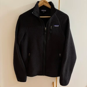 Svart fleecejacka från Patagonia - Svart fleece från Patagonia i storlek S. Jackan har hög krage, tre dragkedjeförsedda fickor och är tillverkad i mjuk fleece av 100% polyester. Perfekt för kalla dagar och har en clean, sportig look med diskret logga på bröstet. Som ny