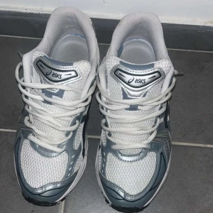 Vita och grå Asics sneakers - Säljer ett par snygga Asics sneakers i vitt och grått med mesh-överdel och detaljer i syntet. Skorna har snörning, platt sula och rund tå. Perfekta för dig som gillar sportig stil och vill ha bekväma skor med cool design.