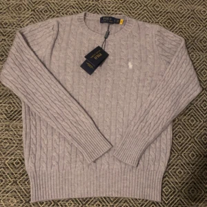 Ljusgrå kabelstickad tröja Polo Ralph Lauren - Kabelstickad tröja från Polo Ralph Lauren i en ljusgrå nyans. Klassisk rund halsringning, långärmad och med diskret broderad logga på bröstet. Tillverkad i mjuk kashmir för en lyxig känsla och perfekt passform.