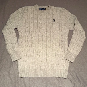 Grå kabelstickad tröja Polo Ralph Lauren - Kabelstickad grå tröja från Polo Ralph Lauren i 100% bomull. Klassisk rund halsringning, ribbade muddar och ikonisk broderad logga på bröstet. Perfekt för lager-på-lager och snygg till jeans eller kjol.