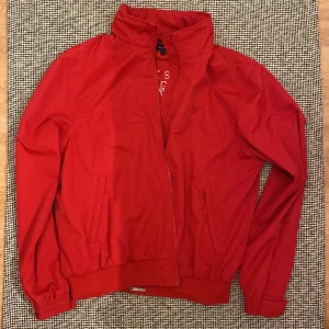 Röd vindjacka från GANT, storlek L - Snygg röd vindjacka från GANT med broderad logga och emblem på insidan. Jackan har hög krage, dragkedja framtill och två fickor med dragkedja. Elastiska muddar vid ärmslut och nederkant ger en sportig vibe. Perfekt för dig som gillar klassisk amerikansk stil.