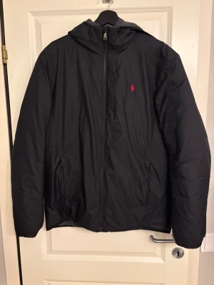 Svart dunjacka från Polo Ralph Lauren - Svart dunjacka från Polo Ralph Lauren med huva och röd logga på bröstet. Jackan har dragkedja framtill, två sidofickor och quiltad insida. Perfekt för kalla dagar och har en clean, stilren look som funkar till det mesta.