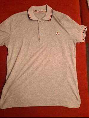 Grå Moncler pikétröja med logga - Snygg grå pikétröja från Moncler med klassisk krage och knappar framtill. Tröjan har Moncler-logga på bröstet och coola ränder i röd, mörkbå och vitt på krage och ärmslut. Tillverkad i mjuk bomull som är skön mot huden.