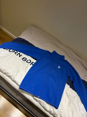 Blå vindjacka från Adidas - Helt oanvänd Snygg blå vindjacka från Adidas med klassisk logga på bröstet. Jackan har hel dragkedja, ståkrage och långa ärmar. Perfekt för dig som gillar sportig stil och vill ha något lätt och bekvämt. Materialet är polyester och jackan har en clean look utan mönster.