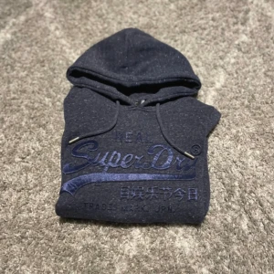 Mörkgblå Superdry hoodie - Snygg mörkgrå hoodie från Superdry med blått broderat logotryck på bröstet och japanska tecken. Tröjan har huva med snörning, känguruficka och långa ärmar. Perfekt för chill dagar och streetstyle.