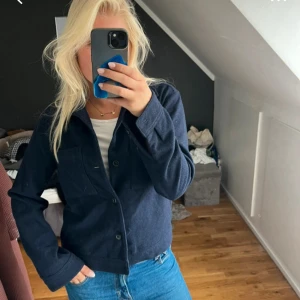 Mörkblå overshirt från Riley - Säljer en stilren mörkblå overshirt från Riley med klassisk krage och två bröstfickor. Jackan har knäppning framtill med svarta knappar och är långärmad. Materialet är mjukt och känns som ull, perfekt för lager-på-lager under höst och vår.