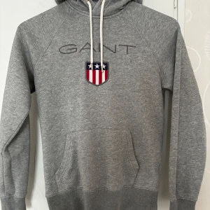 Grå hoodie från GANT  - Grå hoodie från GANT i storlek XS med broderad logga och emblem på bröstet. Tröjan har huva med vita snören, känguruficka och långa ärmar. Materialet är mjuk bomull och polyesterblandning, perfekt för chill dagar. Snyggt broderad detalj med stjärnor och ränder i rött, vitt och blått.