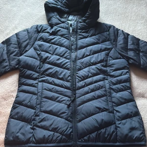 Svart pufferjacka från Tom Tailor - Säljer en svart pufferjacka från Tom Tailor Denim i storlek L. Jackan har huva, dragkedja framtill och snygga quiltade detaljer i ett chevronmönster. Perfekt till våren när det är lite små kallt. 