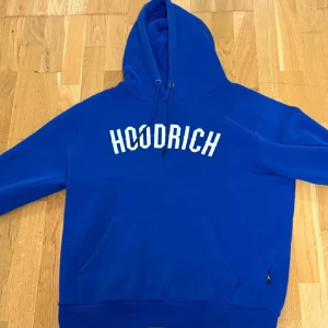 Blå hoodie från Hoodrich med tryck - Snygg blå hoodie från Hoodrich med stor vit logga på bröstet och coolt tryck på ryggen. Tröjan har huva med snörning, känguruficka och långärmade ärmar. Perfekt för dig som gillar streetwear och vill sticka ut med en bold färg.