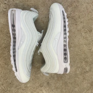 Nike Air Max 97 vita sneakers strl 40 - Nike Air Max 97 sneakers i helvitt med ikonisk vågig design och synlig Air-sula. Skorna har snörning, rund tå och platt sula. Materialet är en mix av syntet och textil, med diskret Nike-logga på sidan och innersulan. Perfekt för dig som gillar clean och sportig stil.