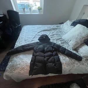 Svart Sail Racing dunjacka med huva - Svart dunjacka från Sail Racing, modell Antarctica Expedition. Jackan har huva med logga, dragkedja framtill och patch på ärmen. Puffad design med snygga paneler och diskreta detaljer. Perfekt för kalla dagar och har en clean look som funkar till streetwear. Har dock ett litet hål vid vänstra armen märker knappast