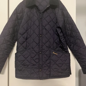Mörkblå quiltad jacka Barbour  - Quiltad jacka från Barbour i mörkblå färg, storlek XL ungdom, den passar mig som är 173. Klassisk design med tryckknappar, två stora fickor och Barbour-logga broderad på fickan. Jackan har lång ärm och krage i kontrastmaterial. Perfekt för dig som gillar stilren och tidlös look. Skick 10/10