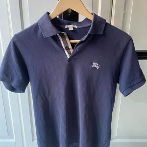 Mörkblå pikétröja från Burberry - Snygg mörkblå pikétröja från Burberry med klassisk krage. Tröjan har Burberrys ikoniska rutiga detalj vid knäppningen. Bra skick. Bara att fråga på