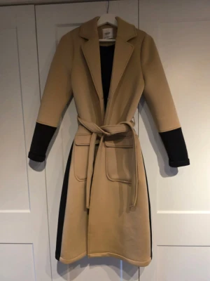 Beige kappa från Summum Woman XS - Snygg beige kappa med svarta detaljer från Summum Woman i storlek XS. Jackan har stora fickor, bälte i midjan och knappar framtill. Långärmad och med klassisk krage, perfekt för dig som gillar stilrena och moderna plagg.